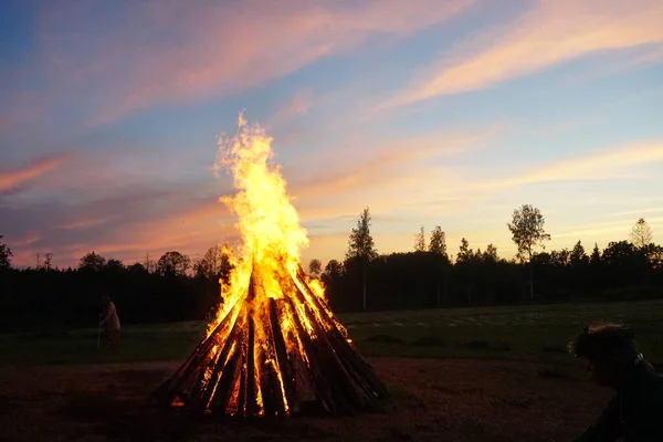 A large bonfire burns at midsummer night, with the sun setting in the background