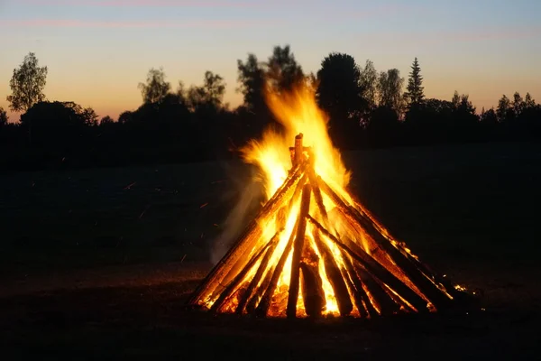 A large bonfire burns at midsummer night, with the sun setting in the background