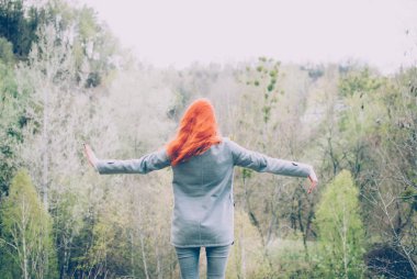 Kızıl saçlı kız tepenin önünde dağ orman kenarında duran ve ellerini yükselterek gri bir ceket giyiyor. Konsept (fikir) özgürlük, rüya, sakinlik, meditasyon ve motivasyon.
