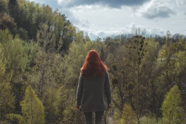 Tepenin önünde dağ orman kenarında bekleyen bir gri ceket giyen longhair Kızıl saçlı kız. Konsept (fikir) özgürlük, hayal, düşünce, meditasyon ve motivasyon. Bulutlu bahar öğleden sonra.