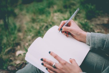 Not defterinde düşünceleri aşağı yazma yeşil bahar çimenlerin üzerinde oturan kız. Solgun elleri ve boş kağıt defter. Konsept (fikir) düşünme, hayal, motivasyon, planlama, öğrenme ve eğitim. Erken bahar öğleden sonra.