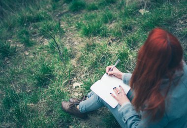 Kızıl saçlı kız aşağı not defterinde düşünceleri yazma yeşil bahar çimenlerin üzerinde oturan gri bir ceket giyiyor. Solgun elleri ve boş kağıt defter. Düşünme, hayal, planlama, öğrenme ve eğitim konsept (fikir). Erken bahar öğleden sonra.
