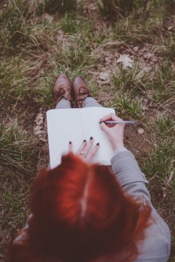 Not defterinde düşünceleri aşağı yazma yeşil bahar çimenlerin üzerinde oturan kızıl saçlı kız. Solgun elleri ve boş kağıt defter. Konsept (fikir) düşünme, hayal, motivasyon, planlama, öğrenme ve eğitim. Erken bahar öğleden sonra.