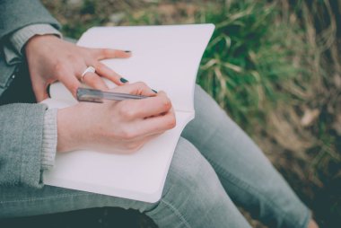 Not defterinde düşünceleri aşağı yazma yeşil bahar çimenlerin üzerinde oturan kızıl saçlı kız. Solgun elleri ve boş kağıt defter. Konsept (fikir) rüya, motivasyon, düşünme, planlama, eğitim.