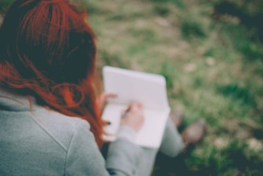 Ufuk arka plan bulanık. Not defterinde yazılı yeşil bahar çimenlerin üzerinde oturan kızıl saçlı kız. Solgun elleri ve boş kağıt defter. Konsept (fikir) rüya, motivasyon, düşünme, planlama, eğitim.