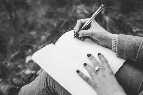 Redhead girl sitting on spring grass writing down her thoughts in notebook. Pale hands and blank paper of notebook. Concept (idea) of thinking, dreaming, motivation, planning, studying. Black and white