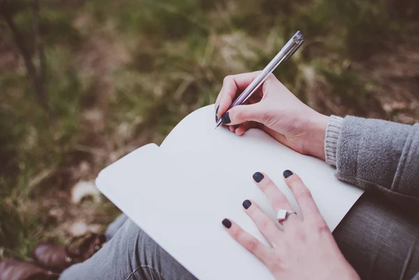 Girl sitting on a green spring grass writing down her thoughts in the notebook. Pale hands and blank paper of the notebook. Concept (idea) of thinking, dreaming, motivation, planning, learning, and studying. Early spring afternoon.