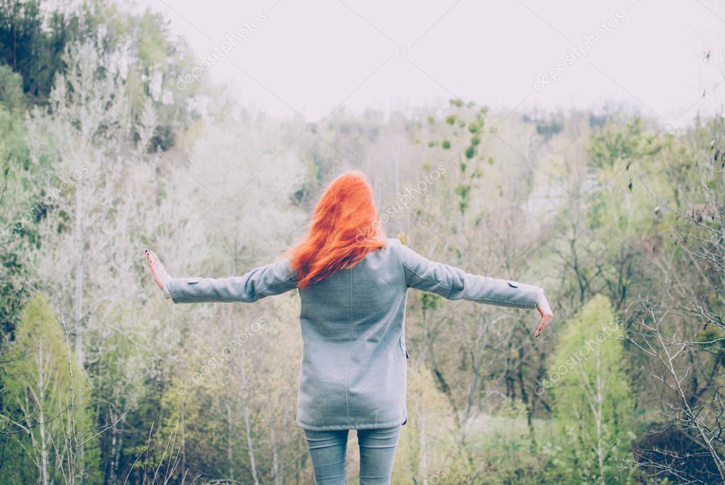 Redhead girl wearing a grey coat standing on the edge of the hill in front of the forest of the mountain and raising her hands. Concept (idea) of freedom, dreaming, calmness, meditation, and motivation.