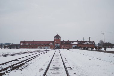 Oswiecim, Poland - February 16, 2018: Railway tracks of Auschwitz concentration camp, the famous watchtower, and visitors clipart