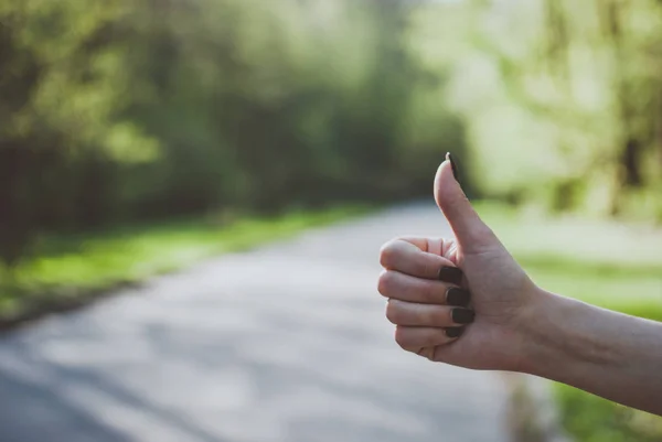 Girl making a thumps up (like) gesture against sunny green background. Concept of hitchhiking, traveling, tourism, and adventures