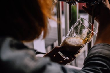 Kız bir bardak bira zanaat bira barda koyu bira döküyor. Yerel pub. Parlak bira arka plan üzerinde musluklar. Parlak güneşli öğleden sonra. Bir omuz bakmak