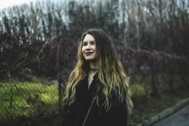 Kameraya gülümseyen güzel uzun saçlı kız. Bulanık cadde ve park ağaçlar bir arka plan üzerinde. Renkli kıvırcık saçlı güzel bir model'ın omuzlarında azalan. 