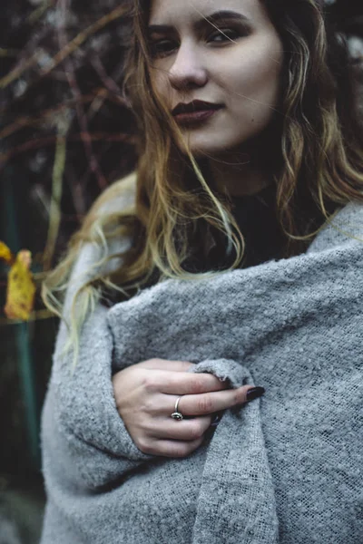 Mysterious look of a charming black-eyed caucasian model covered in a warm gray blanket. Woman standing by dry bushes near the fence of a neighborhood. Dreamy romantic girl.