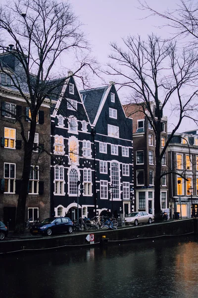 Tradycyjny murowany holenderski domy w Amsterdamie, Holandia. Wąskie czarne i pastelowe ściany elewacji z wysokimi oknami na zimne wieczorne niebo. Światła okienne odbijane w wodzie kanału — Zdjęcie stockowe