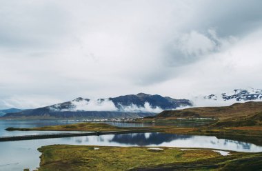 Gri ağır gökyüzü altında renkli İzlanda manzaralar. Yeşil ıslak otlar, güzel göl ve karlı tepelerinin tepesi göl suyuna yansır. Kirkjufellsfoss, Snaefellsnes yarımadası.