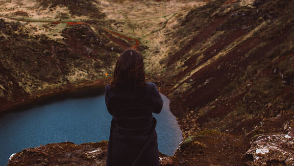 Brunette girl in the gray coat standing on the edge of the red rock surrounding the lake of Kerid (Kerith) located in a volcanic crater. Her hair blowing in wind. Dreaming and traveling concept. Look from behind.