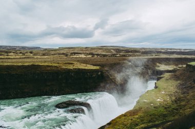 İzlanda'da Gullfoss şelalesi yeşil alanlar, siyah kayalar ve turkuaz su. Havada uçan sıçrama bulutları
