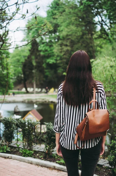 Girl in a striped white and black shirt with a leather bag standing in front of the lake. Look from behind