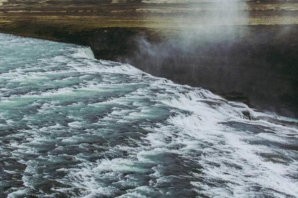 Dreamy Icelandic landscape with blue water with white foam flowing over black stones and falling from edge of the waterfall — Stock Photo, Image