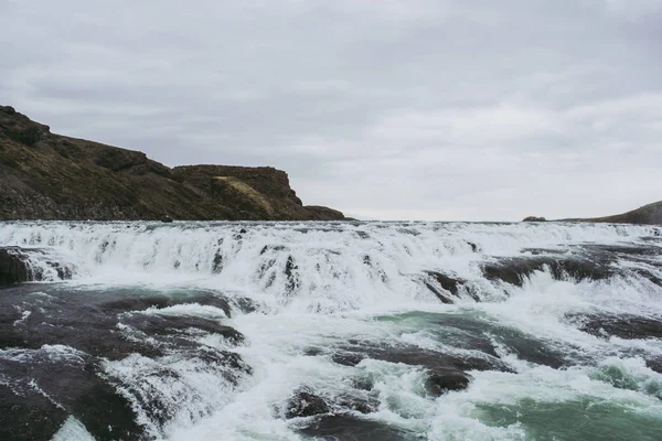 Kaszkád vízesés Izlandon. Fehér kifröccsenés, türkiz víz, fekete szikla a szürke ég alatt. Tavaszi — Stock Fotó