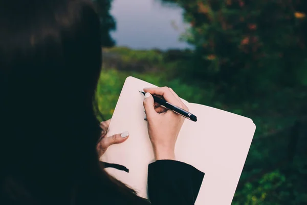 Dziewczyna piśmie w dół jej myśli w Notatniku przeciwko maleńkiej niewyraźne łabędzia domu w środku stawu w parku. Blade strony i kolorowe tło. — Zdjęcie stockowe