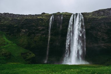 İzlanda Ünlü Seljalandsfoss şelale. Doymuş yeşil çimenler, soluk gri kayalar ve şiddetli yağmurlu gökyüzü