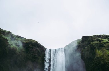 İzlanda'da yeşil tepeler arasında Skogarfoss şelale düşen su. Gri bulutlu gökyüzü ve beyaz sıçramalar.