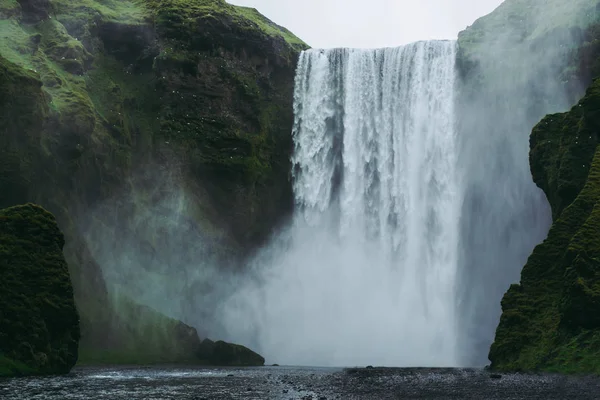 アイスランドの雄大なスコガルフォスの滝。苔で覆われた緑の丘に向かって飛び回る白と緑の水しぶき. — ストック写真
