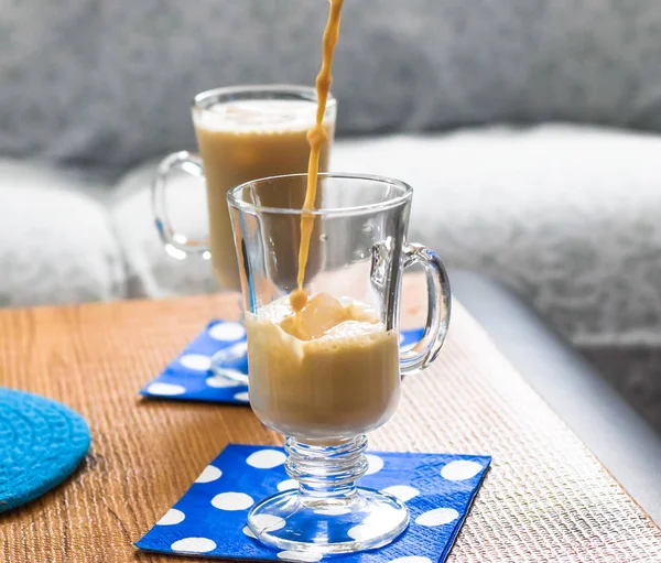 Pouring iced coffee from top in a glass mug with ice cubes, cold coffee summer drink on wood surface with blue napkins, glass of ice coffee