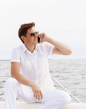 Yakışıklı adam bir yatın güvertesinde oturan ve bir telefonda konuşurken güneş gözlüğü. Yaz aylarında konsept.