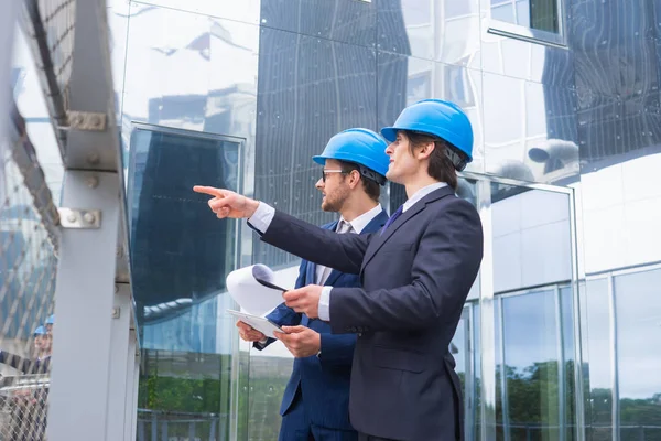 Real estate developers in helmets. New office construction. Confident business men and architect talking in front of modern office building. Businessman and his colleague have conversation.