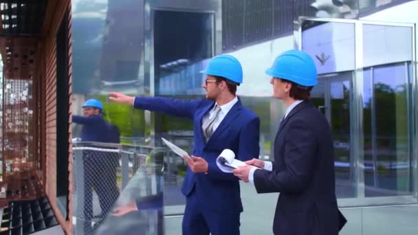 ヘルメットの不動産開発者 新しいオフィス建設 現代のオフィスビルの前で話す自信のあるビジネスマンや建築家 ビジネスマンと彼の同僚が会話を持っている — ストック動画