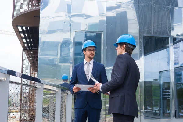 Real estate developers in helmets. New office construction. Confident business men and architect talking in front of modern office building. Businessman and his colleague have conversation.