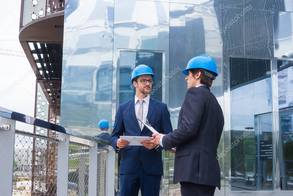 Real estate developers in helmets. New office construction. Confident business men and architect talking in front of modern office building. Businessman and his colleague have conversation.