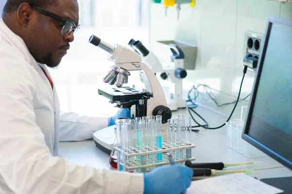 Um afro-americano a trabalhar no laboratório. Médico cientista a fazer pesquisa médica. Ferramentas de laboratório: microscópio, tubos de ensaio, equipamento. Biotecnologia, química, ciência, experiências e cuidados de saúde. — Fotografia de Stock