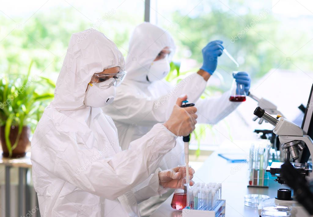 Scientists in protection suits and masks working in research lab using laboratory equipment: microscopes, test tubes. Medicine, infection and vaccine discovery concept.