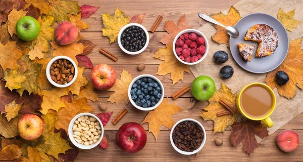 Fondo de otoño estacional. Marco de hojas de arce y un pastel, bayas, pasas, manzanas, frutas, café y nueces sobre fondo de madera . — Foto de Stock