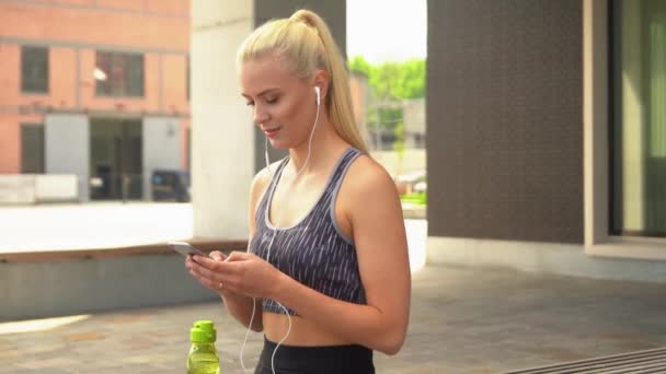 Junges, attraktives und sportliches blondes Mädchen in Sportbekleidung, das der Musik lauscht und sich im Freien entspannen kann. Gesundheit, Sport, Fitness und Lifestyle. — Stockvideo