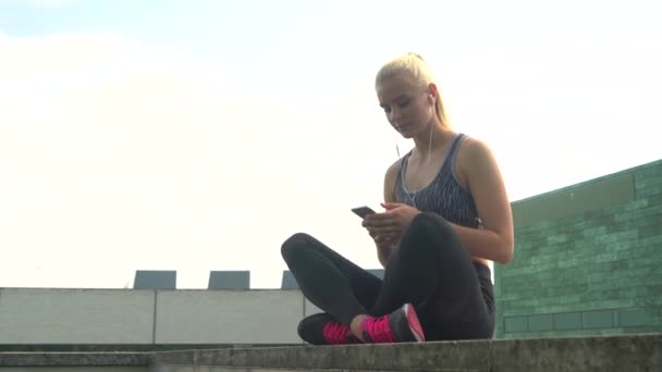 Jong, aantrekkelijk en sportief blond meisje in Sportswear luisteren naar de muziek en ontspannen buiten. Gezondheidszorg, sport, fitness en lifestyle. — Stockvideo