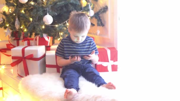 Menino atraente assistindo desenhos animados na frente da árvore de Natal com bolas e luzes. Criança se preparando para o Ano Novo em casa . — Vídeo de Stock