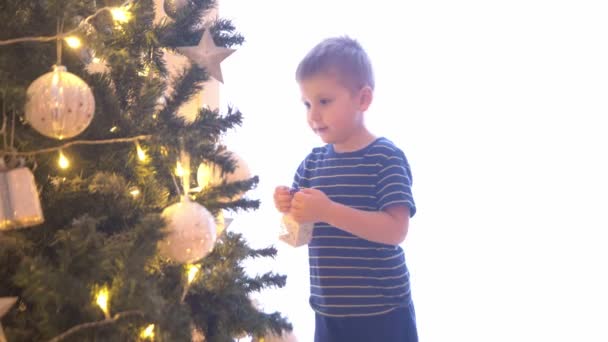 Attraktive kleine Junge schmücken Weihnachtsbaum mit Kugeln und Lichtern. Kind bereitet sich zu Hause auf das neue Jahr vor. — Stockvideo