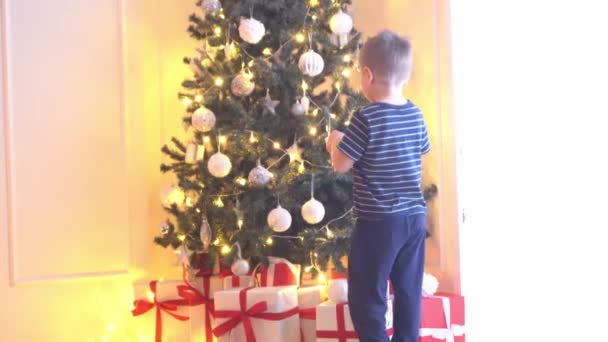Привабливий маленький хлопчик прикрашає ялинку кулями та вогниками. Хлопчик готується до нового року вдома.. — стокове відео