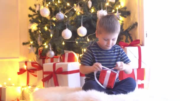Ελκυστικό αγοράκι που ξετυλίγει χριστουγεννιάτικα δώρα. Το παιδί που ανοίγει το νέο έτος παρουσιάζει στο σπίτι. — Αρχείο Βίντεο