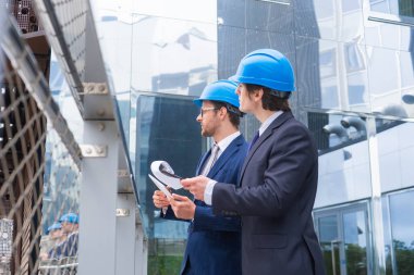 Kasklı emlak çırakları. Yeni ofis inşaatı. Kendine güvenen iş adamları ve mimar modern ofis binası önünde konuşuyor.