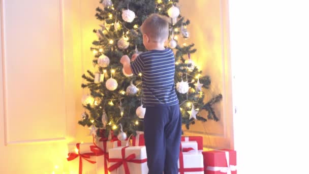 Ελκυστικό αγοράκι που στολίζει χριστουγεννιάτικο δέντρο με μπάλες και φώτα. Παιδί ετοιμάζεται για το νέο έτος στο σπίτι. — Αρχείο Βίντεο