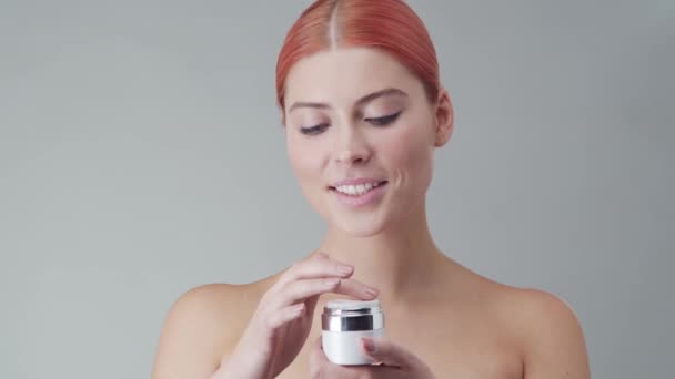 Retrato de estudio de mujer pelirroja joven, hermosa y natural aplicando crema para el cuidado de la piel. Levantamiento facial, cosméticos y maquillaje . — Vídeos de Stock