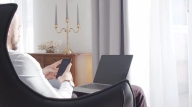 Evde çalışan sakallı iş adamı koltukta oturup bilgisayar teknolojisi kullanıyor. İş, serbest ticaret, yatırımlar ve borsa kavramları.