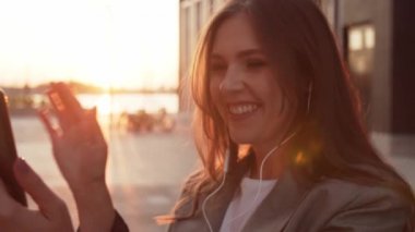 Dışarıda bankta oturan çekici genç bir iş kadını. Video kaset için akıllı telefon kullanan bir kız. Sosyal uzaklık kavramı. Gün batımı ışığı.
