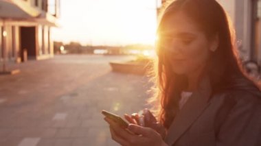 Genç çekici iş kadını dışarıda bankta oturuyor ve akıllı telefon kullanıyor. Gün batımı ışığı.
