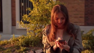 Genç çekici iş kadını dışarıda bankta oturuyor ve akıllı telefon kullanıyor. Gün batımı ışığı.